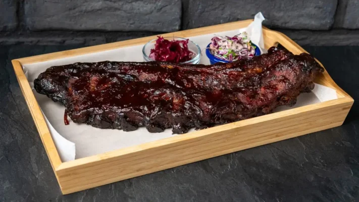 Smoked ribs served on wood plate with coleslaw salad on black brick background